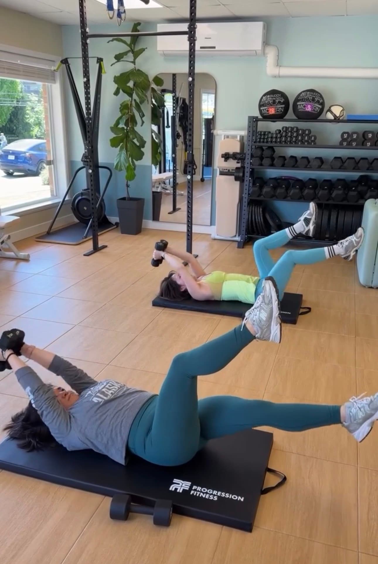 Two women lying on mats in a gym, performing core exercises with dumbbells.
