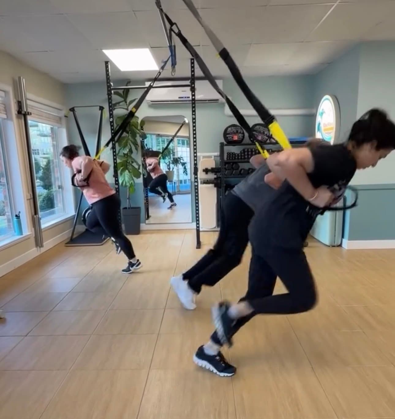 Three women in a fitness studio performing dynamic forward leans using TRX suspension straps.