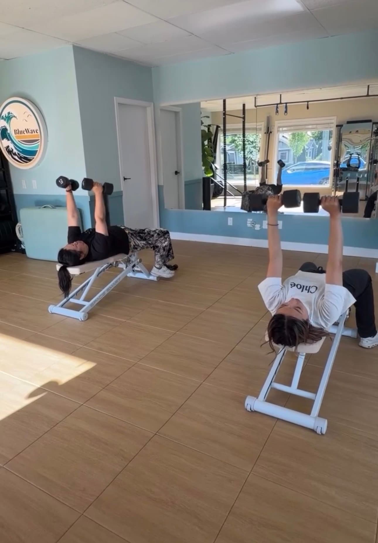 Two women performing dumbbell chest presses on weight benches in a bright fitness studio.