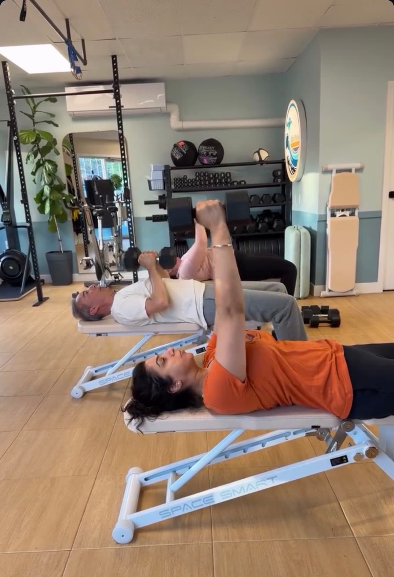 Three people perform dumbbell chest presses on white weight benches in a bright gym.
