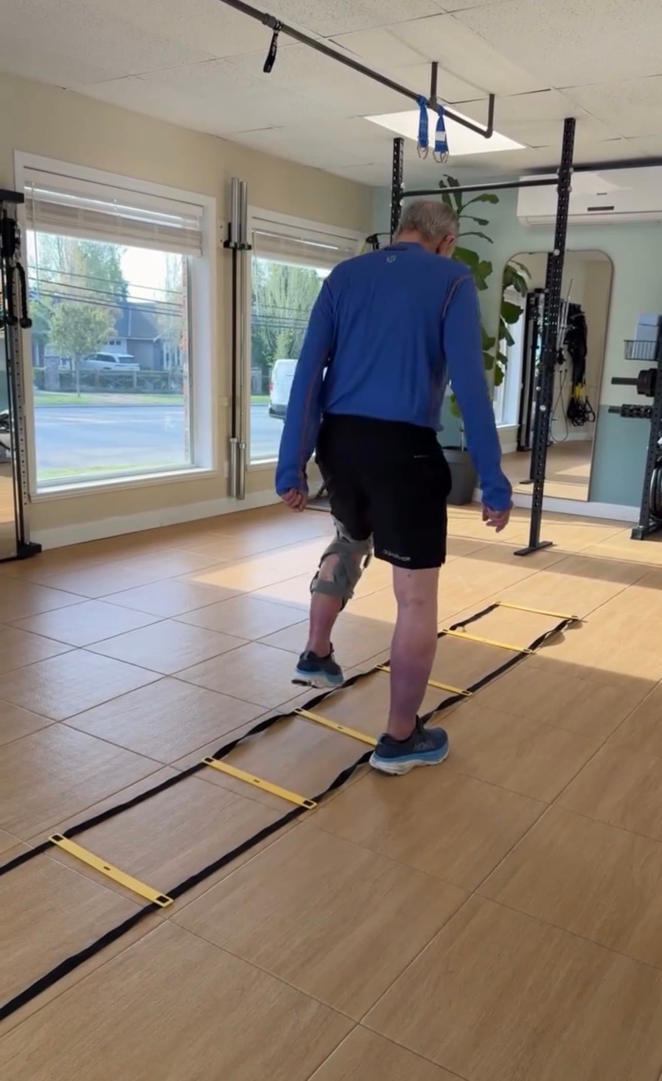 Man with a knee brace steps through a yellow agility ladder in a sunlit gym.