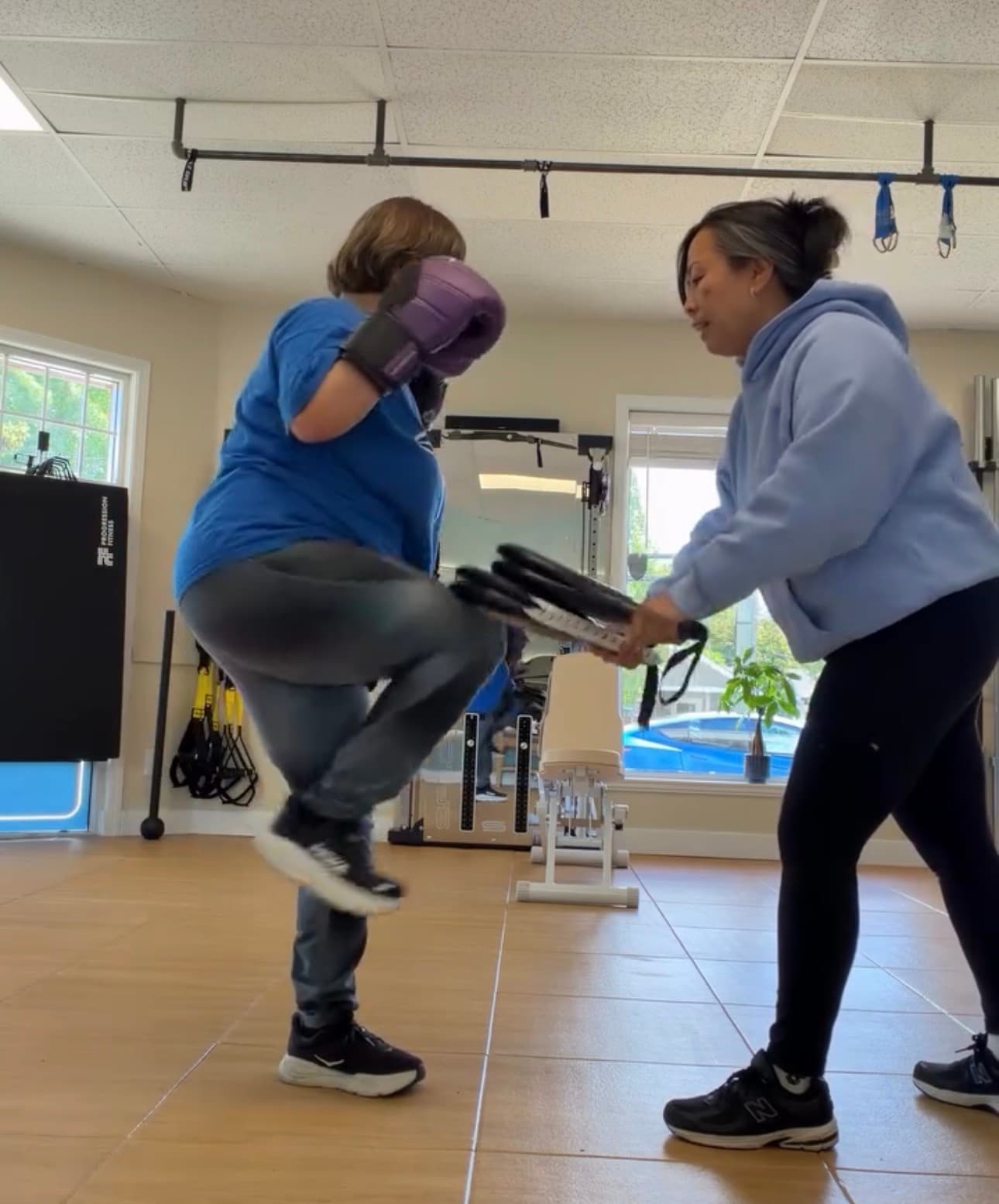 Woman wearing purple boxing gloves performs a knee strike on a pad held by a trainer.