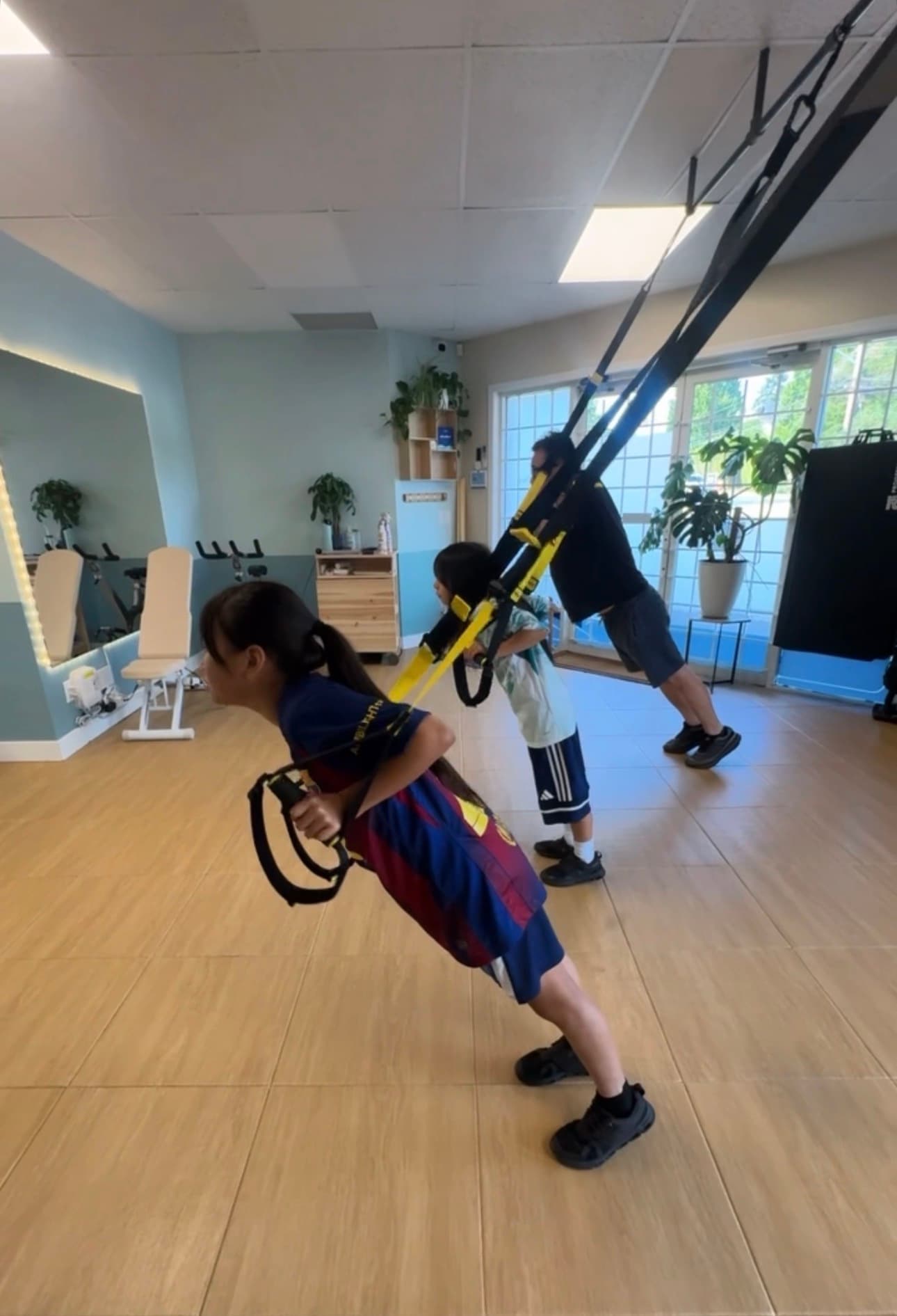 Two children and an adult lean forward using TRX suspension straps in a bright gym.