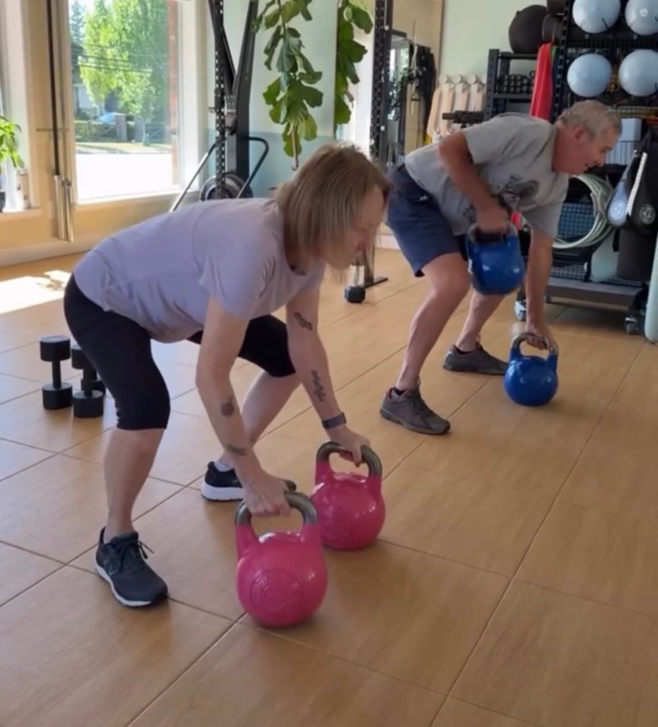 Man and woman perform kettlebell rows in a bright gym with pink and blue weights.