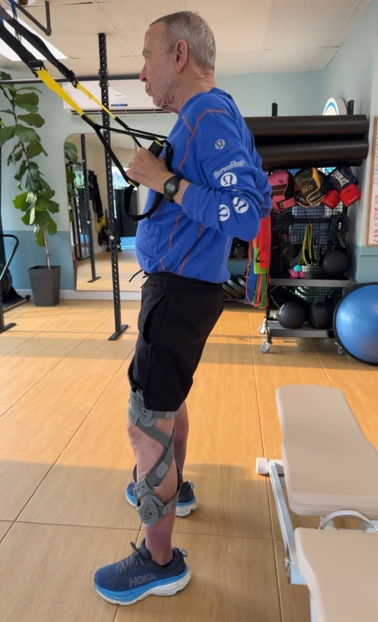 Elderly man wearing a knee brace uses suspension straps for exercise in a gym.