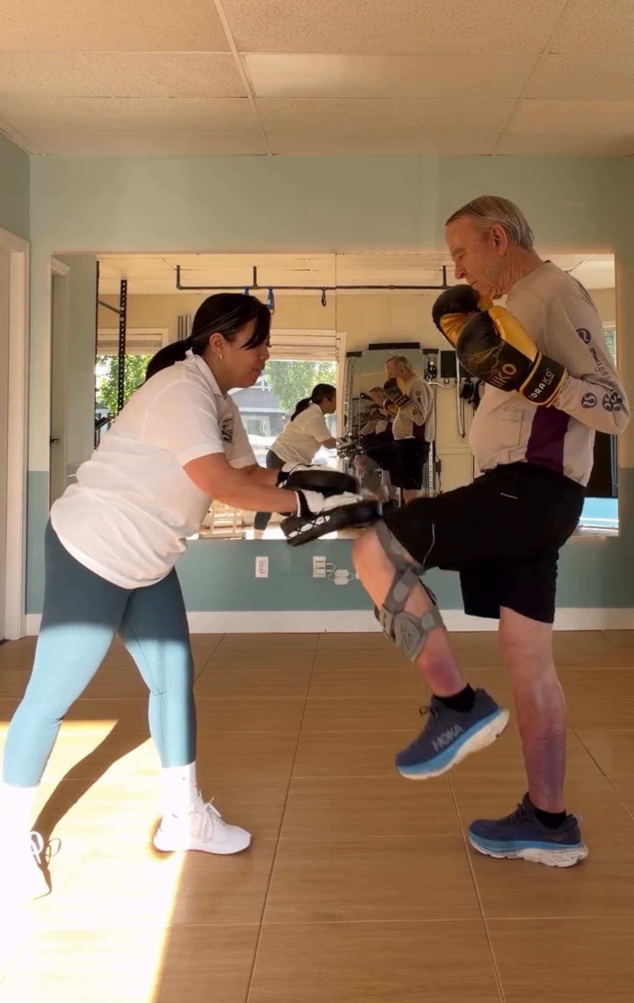 Elderly man wearing a knee brace practices knee strikes against pads held by a trainer.