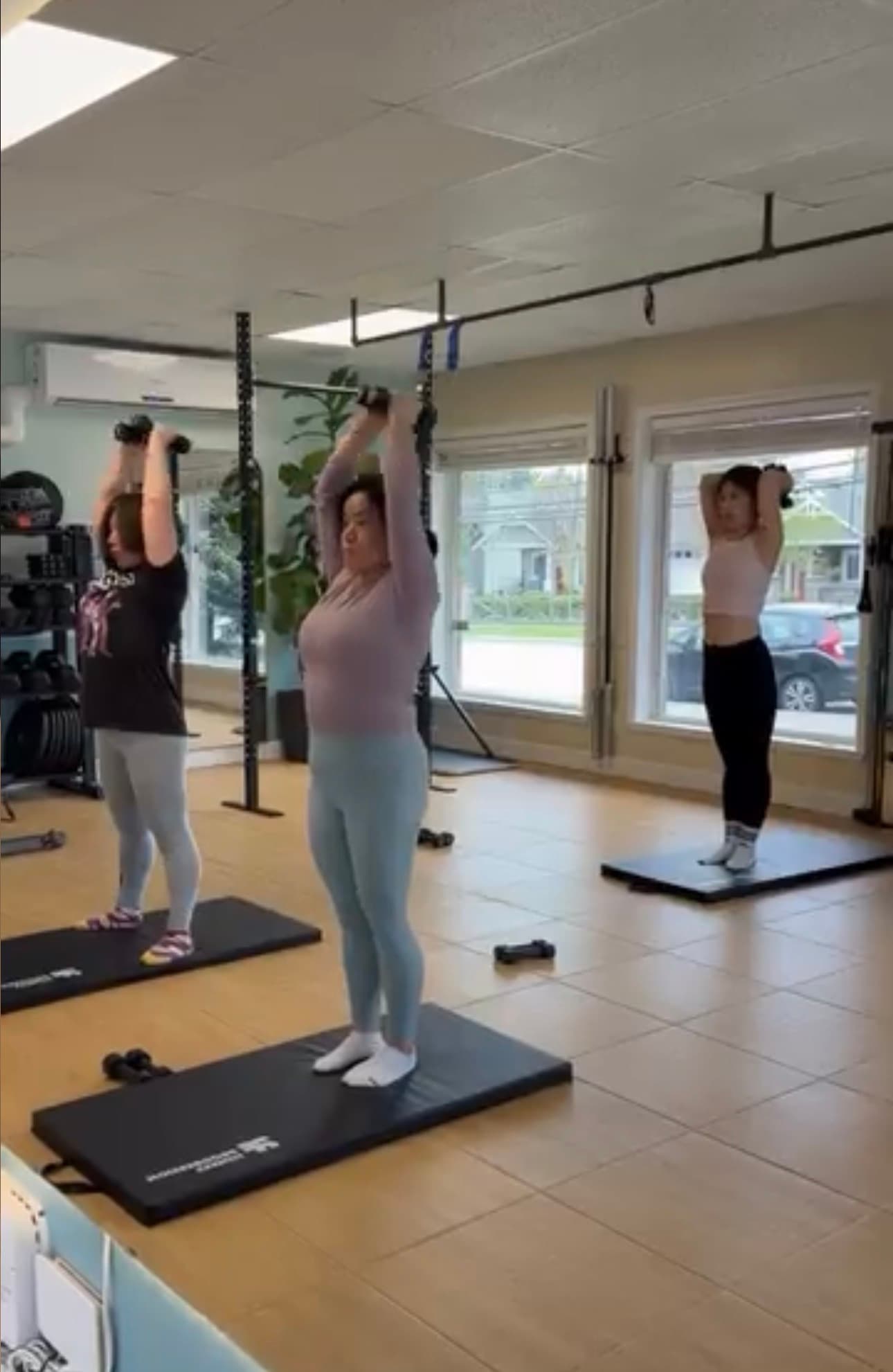 Three women standing on mats in a gym perform overhead tricep extensions using dumbbells.