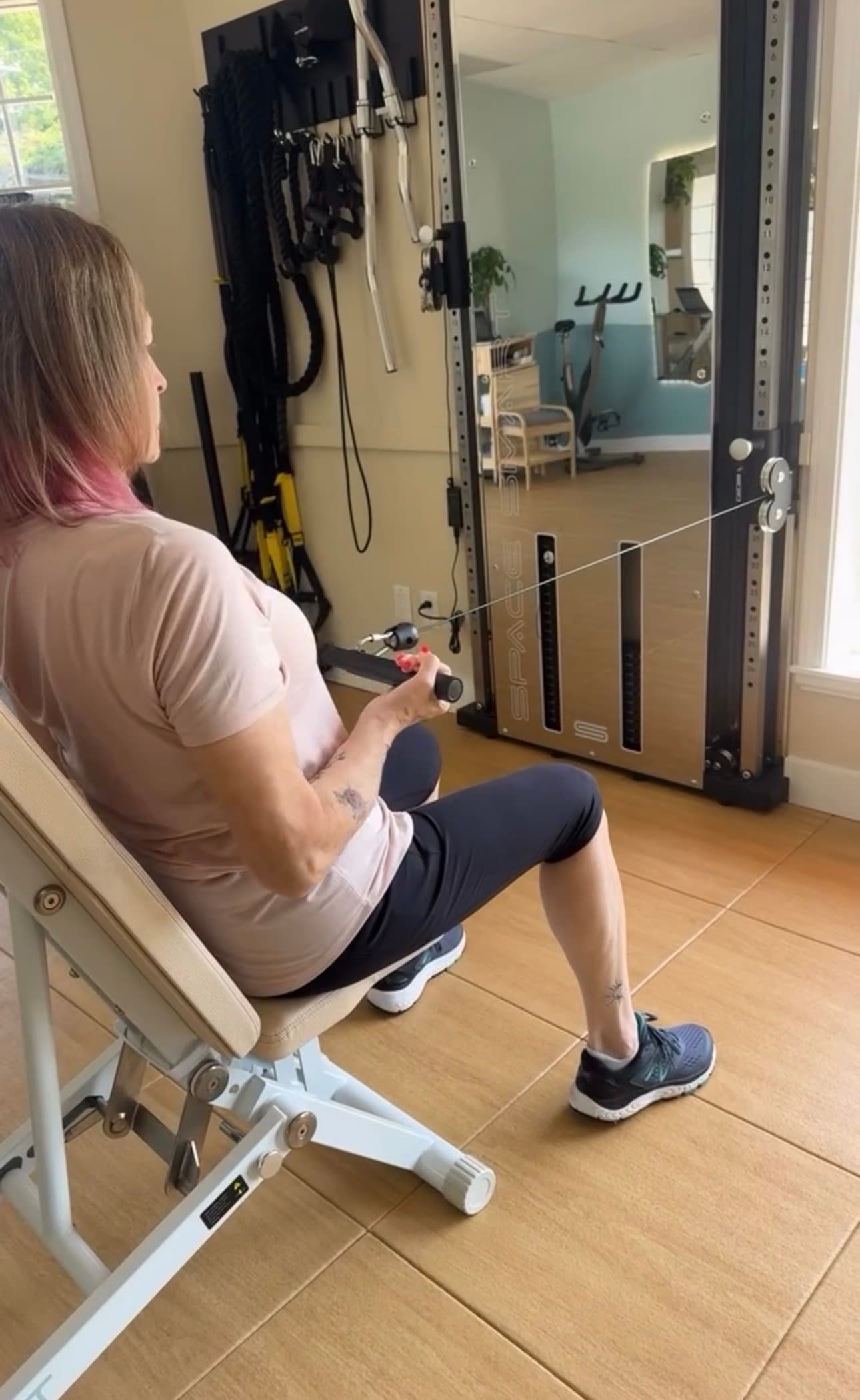 Woman with pink hair performs seated cable rows on a weight bench in a home gym.