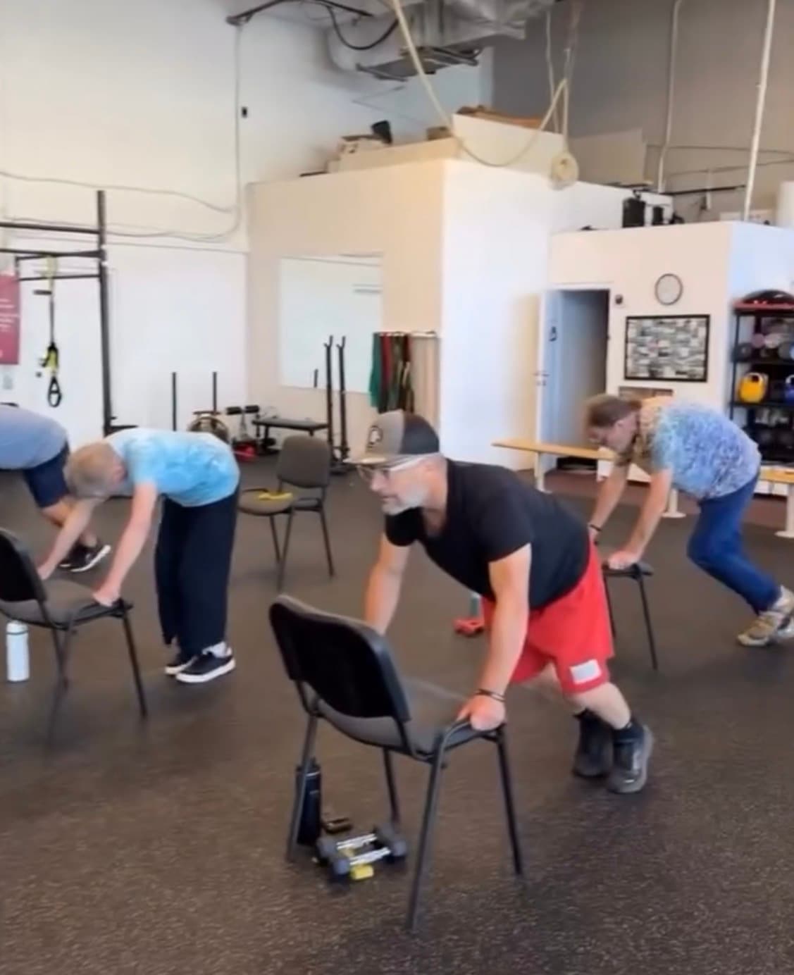 Men in a gym performing mountain climber exercises while leaning on black chairs for support.