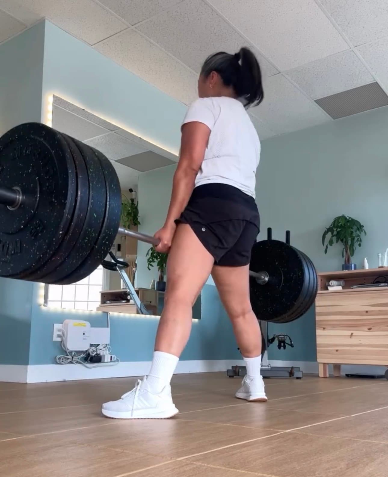 Woman in athletic wear performing a heavy barbell deadlift in a bright home gym.