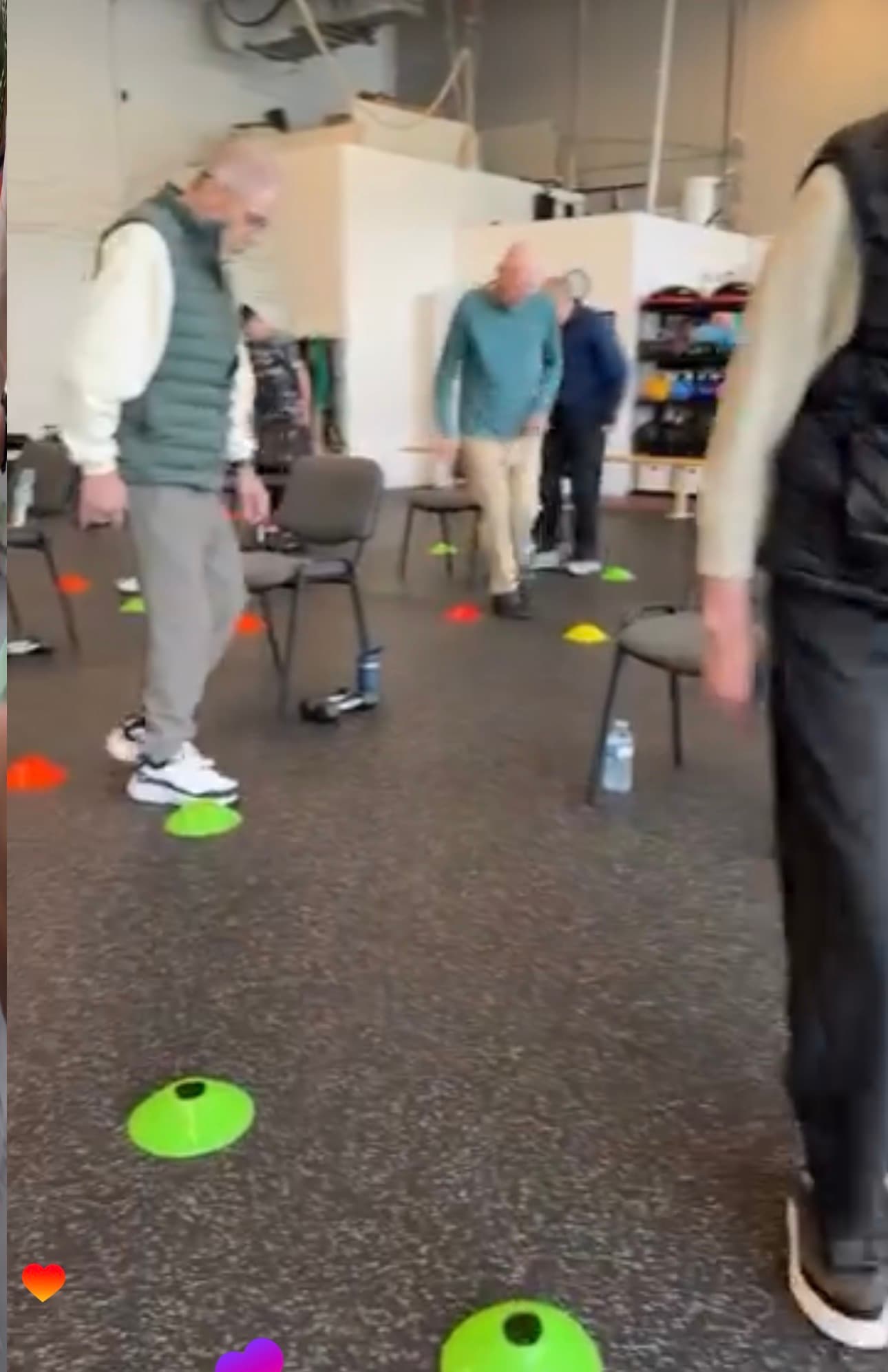 Older men perform agility exercises using colorful cones and chairs in a bright indoor gym.