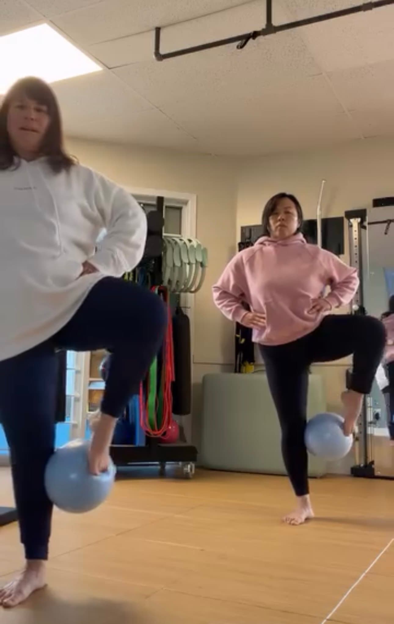 Two women in hoodies balance on one leg using small blue exercise balls.