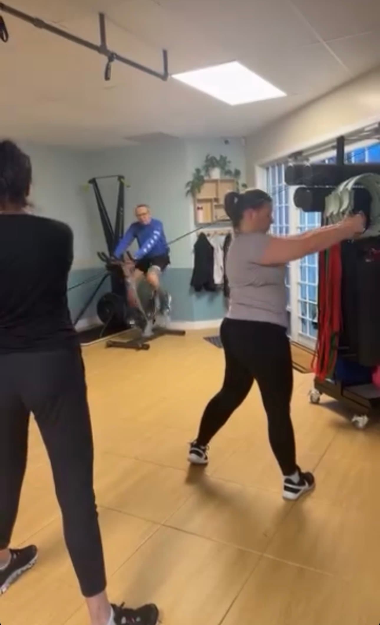 Woman performs cable pulls while a man rides a stationary bike in a gym.
