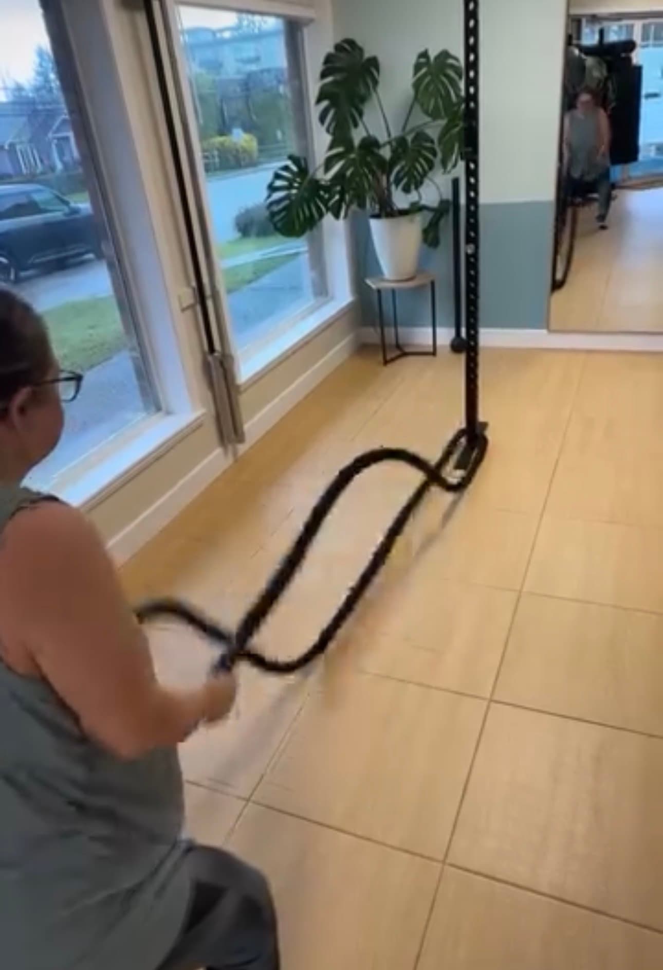 Woman performing battle rope waves in a bright home gym with a large monstera plant.