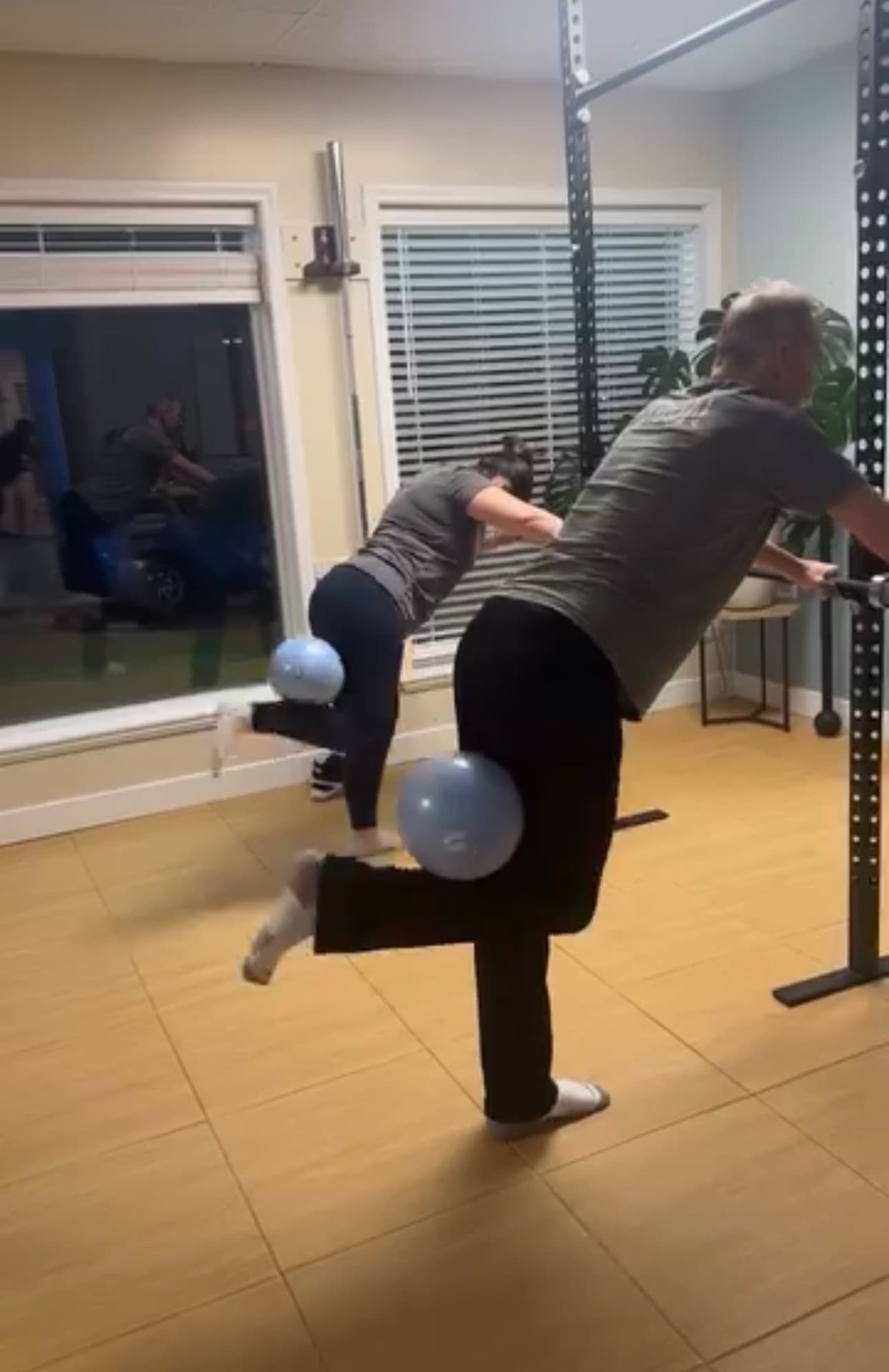 Two people perform leg curls with blue exercise balls behind their knees in a gym.