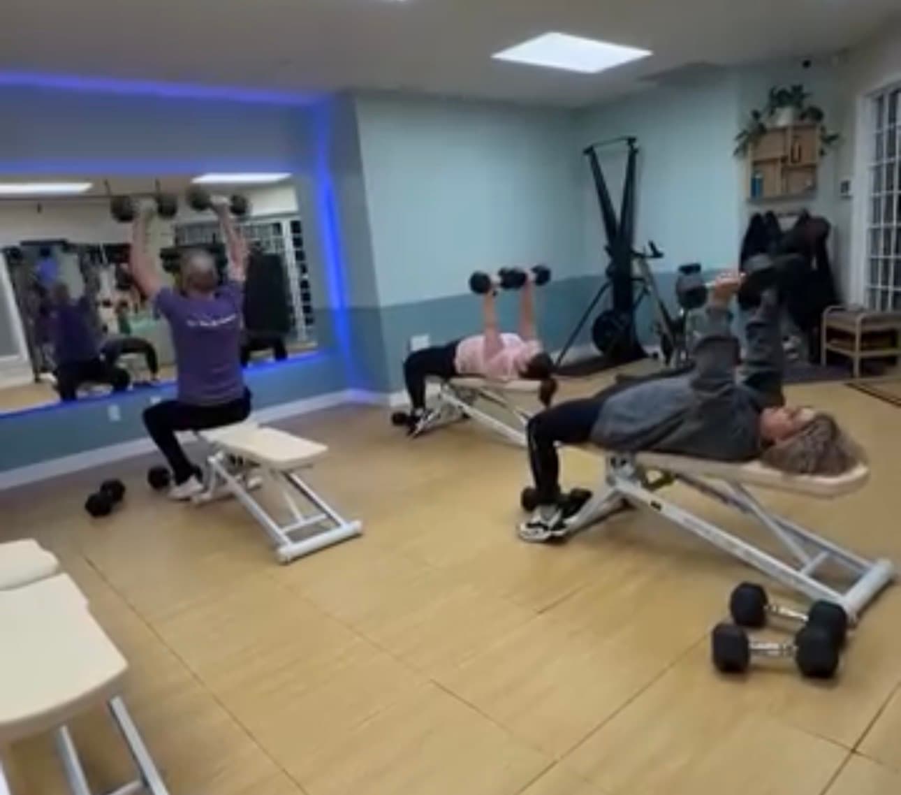 Three people performing dumbbell exercises on weight benches in a brightly lit fitness studio.