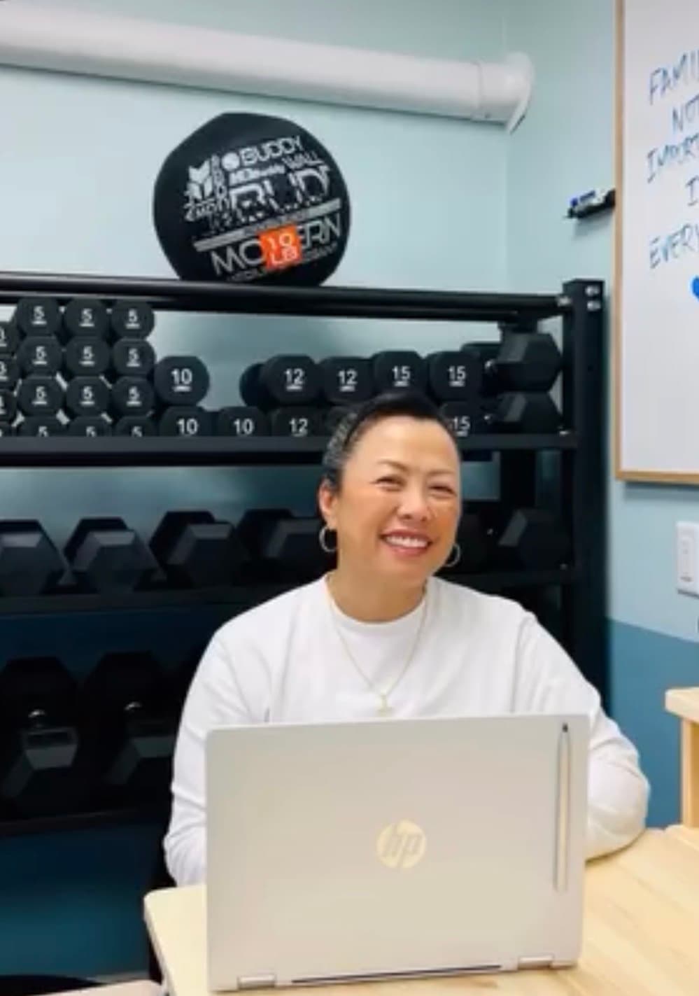 Smiling woman at a laptop in a gym with a rack of dumbbells behind her.