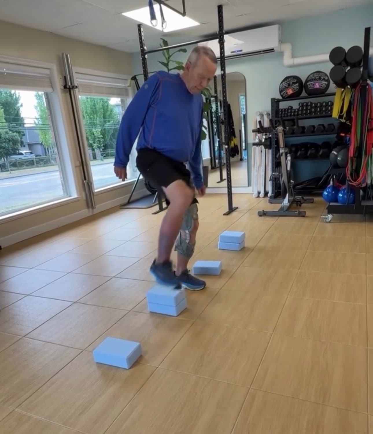 Senior man wearing a knee brace steps over blue foam blocks in a fitness studio.