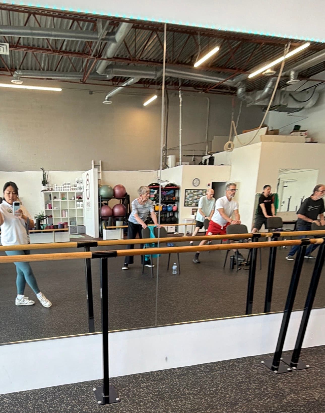 Seniors participate in a fitness class using chairs for balance in a mirrored gym.