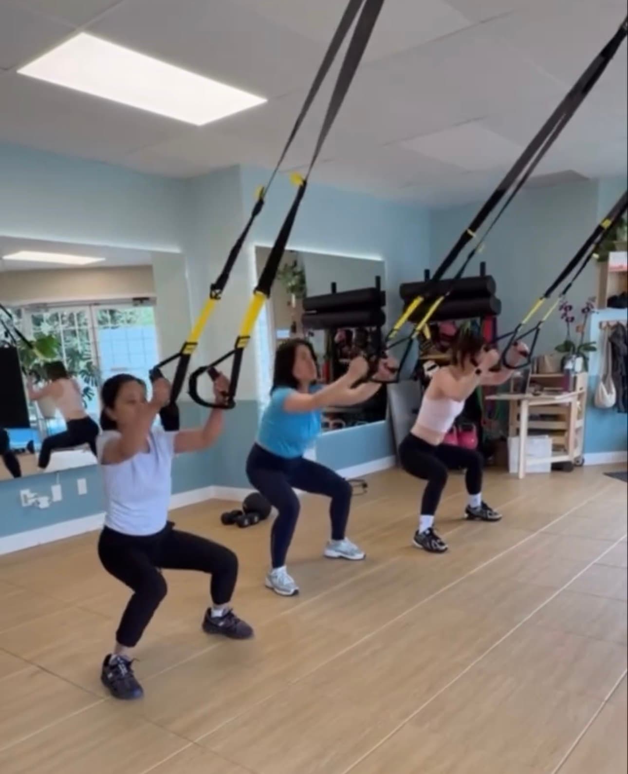 Three women performing squats using TRX suspension trainers in a bright fitness studio.