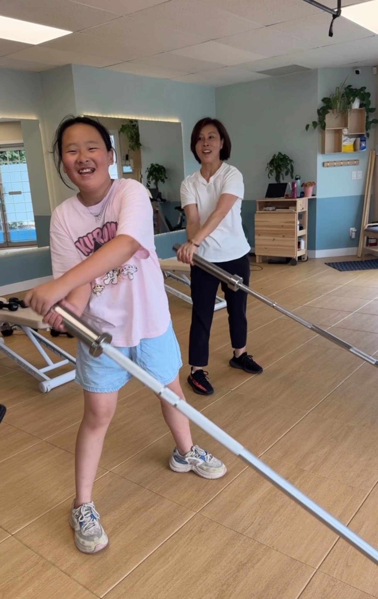 A laughing girl and woman perform rotational exercises with metal bars in a bright studio.