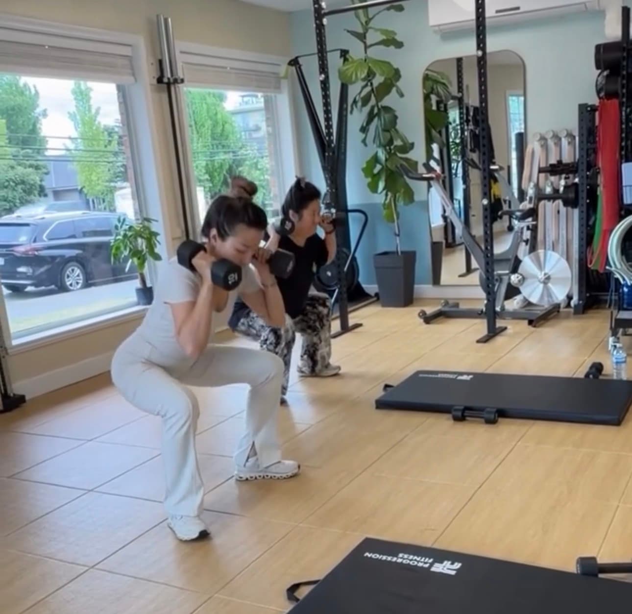Two women performing weighted squats with dumbbells in a bright, well-equipped home gym studio.