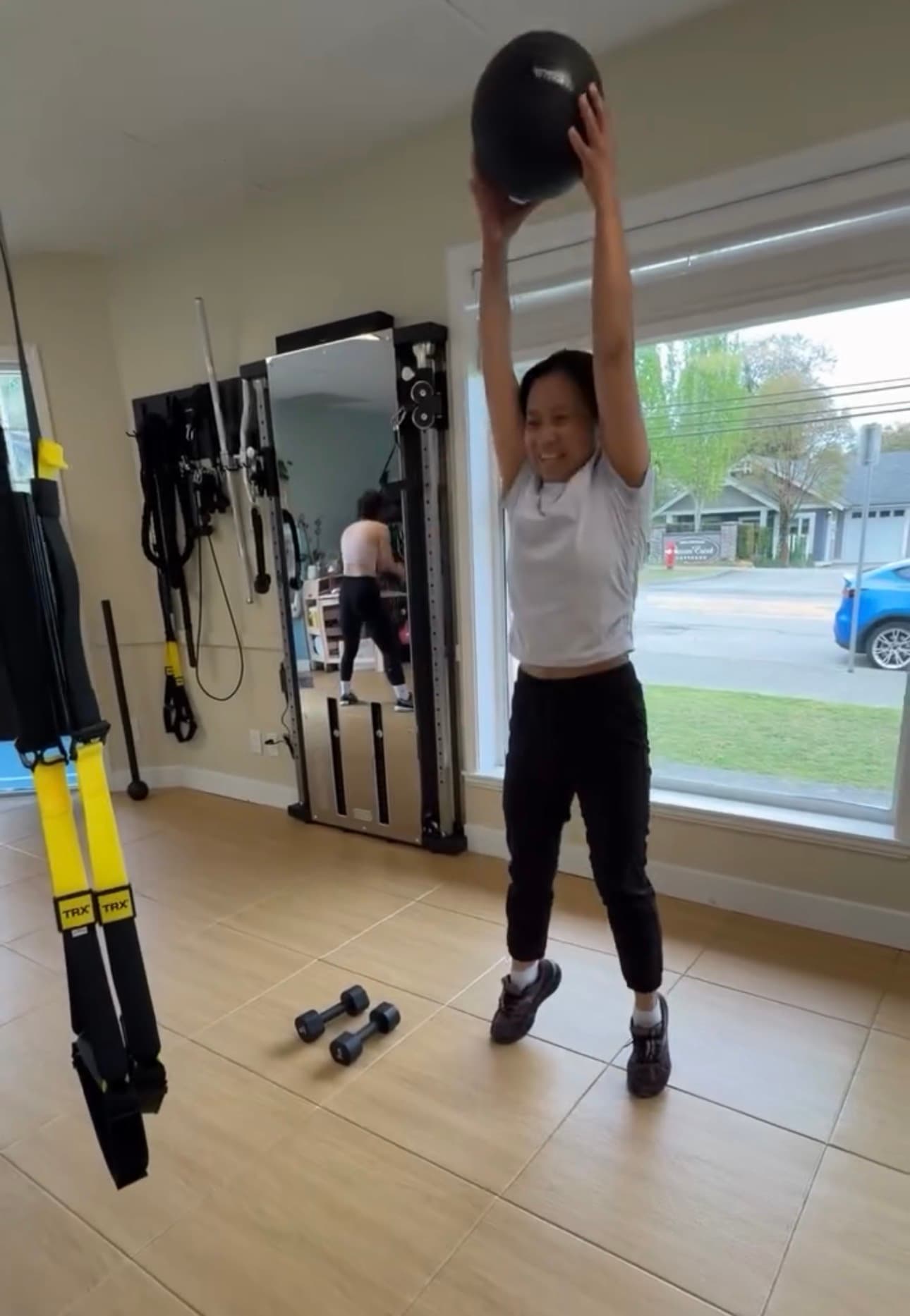 Woman jumping while holding a black medicine ball overhead in a well-lit home gym.
