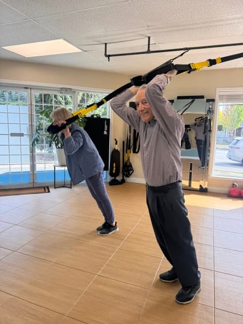 Senior man and woman exercising with TRX suspension straps in a bright fitness studio.