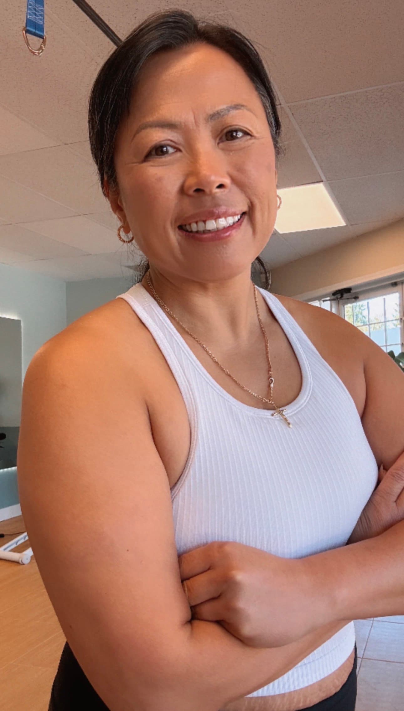 Smiling woman with dark hair wearing a white ribbed tank top and gold cross necklace.